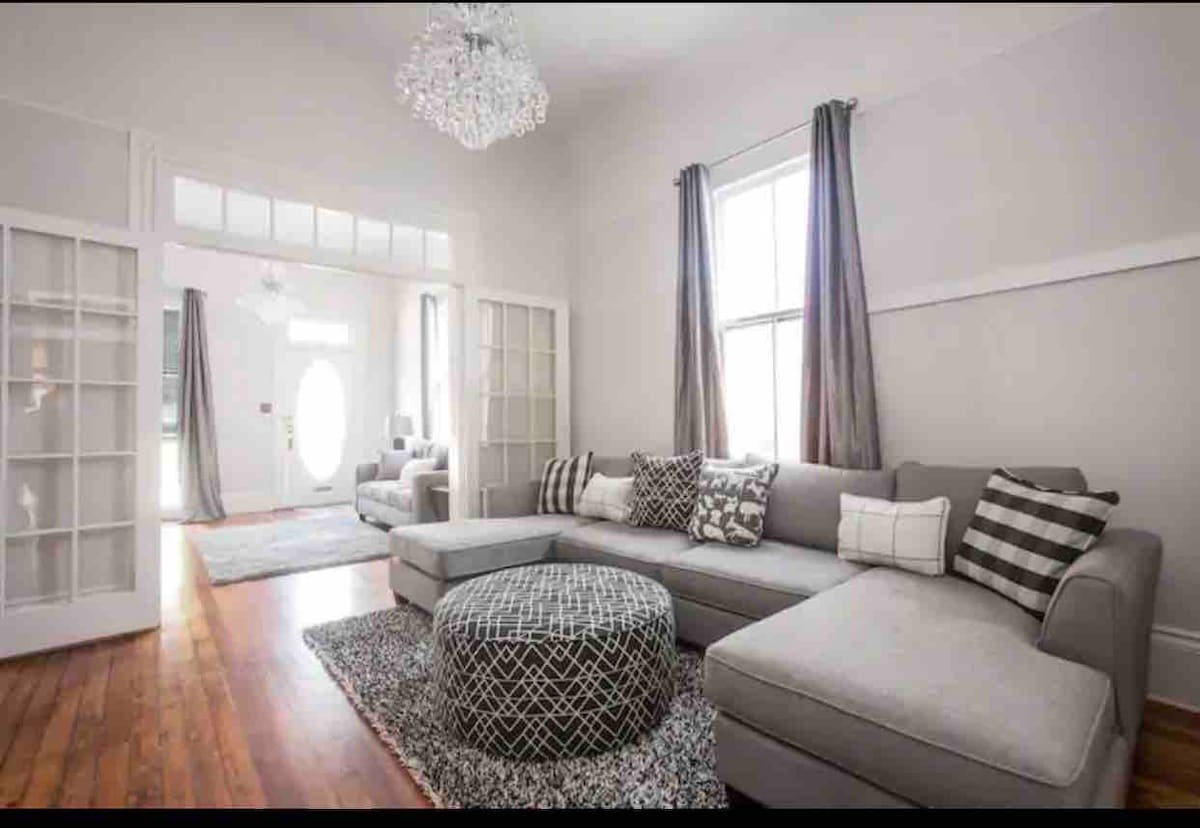 A spacious living area is presented, featuring a large sectional sofa adorned with patterned throw pillows. A textured area rug anchors the seating arrangement. Natural light enters through tall windows, accented by sheer drapes, and a modern chandelier hangs overhead.