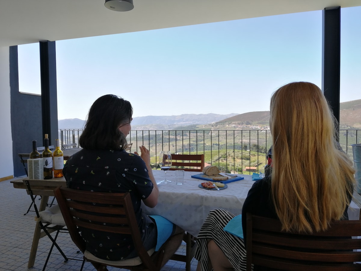 A spacious terrace is featured, offering a panoramic view of the surrounding hills and valleys. Two guests sit at a table adorned with a light tablecloth and various items, while bottles of wine are positioned nearby. The clear sky enhances the view of the picturesque landscape.