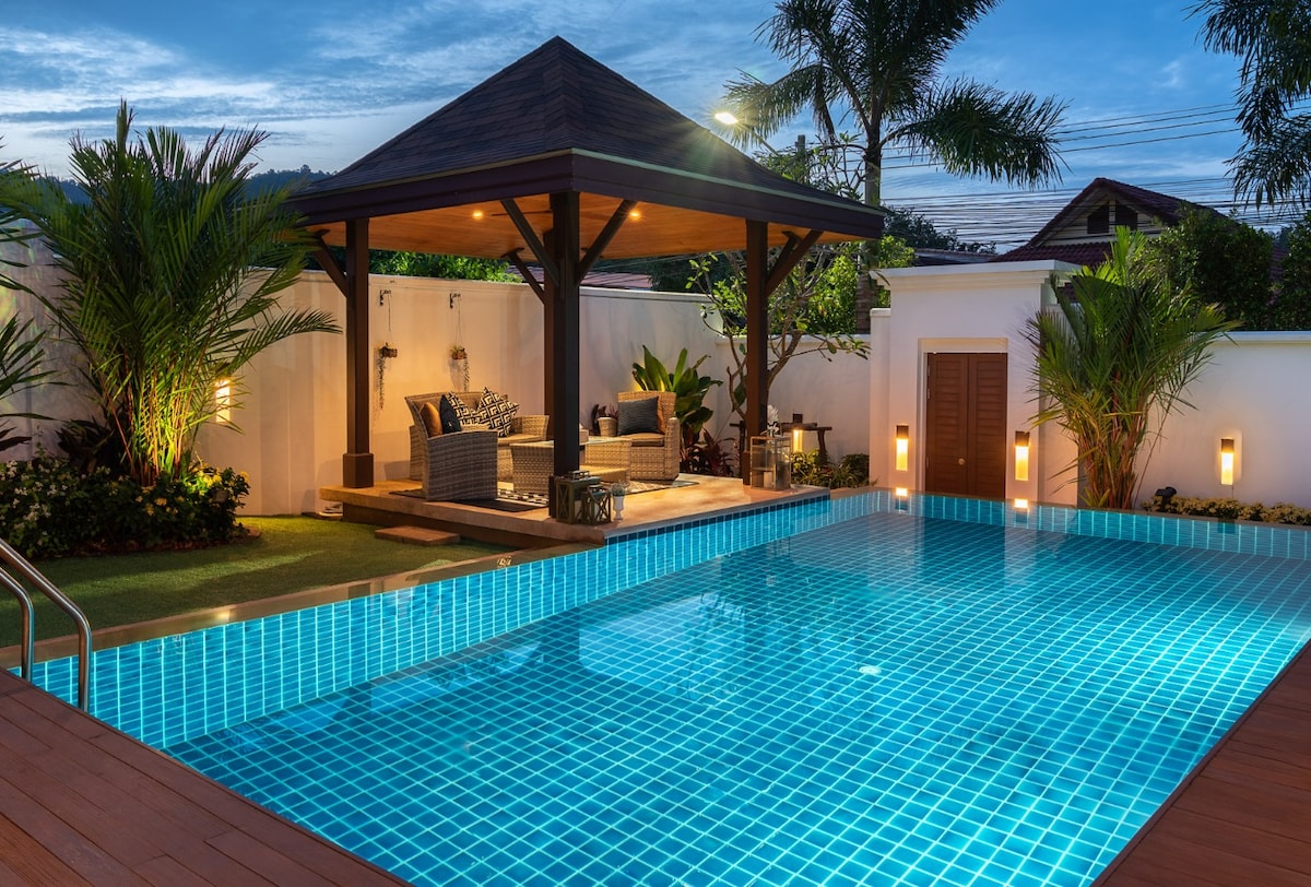 A private swimming pool is surrounded by tropical plants, illuminated by soft lights along the walls. A shaded terrace features comfortable seating and a table, ideal for relaxation. The pool's turquoise tiles reflect the evening sky, creating a serene atmosphere.