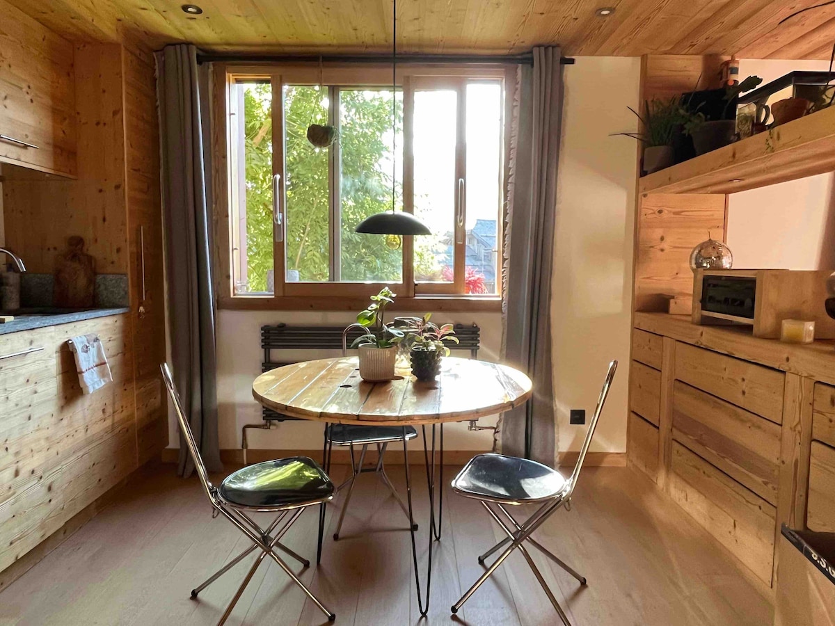 A cozy dining area features a round wooden table surrounded by two metallic chairs. Natural light floods the space through large windows, highlighting the warm wooden paneling. A small plant sits at the center, complemented by understated kitchen cabinetry in the background.