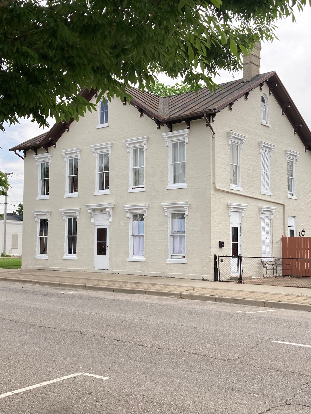 The historic building features a light-colored façade with decorative trim and multiple large windows. A white door is centrally located on the front. A fence outlines the property, providing a sense of boundary along the street.