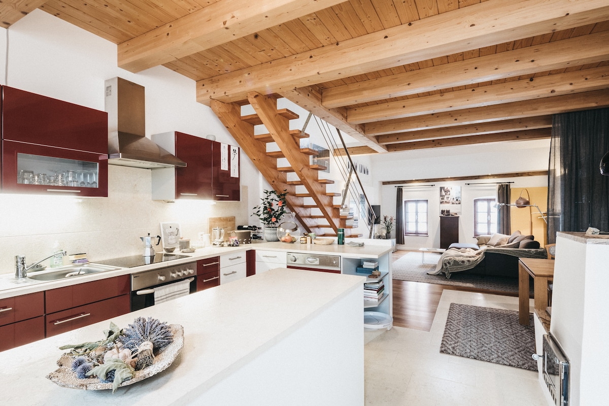 A spacious kitchen area features modern red cabinetry and a large island with a decorative bowl of dried flowers. A wooden staircase leads to the upper level. The living area is visible in the background, showcasing open spaces with comfortable seating and natural light streaming through large windows.