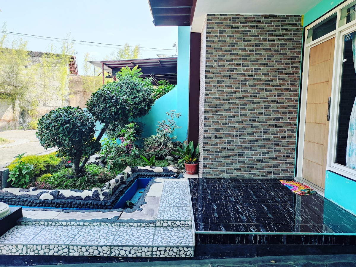 The exterior features a neatly landscaped garden with lush greenery, colorful plants, and a small water feature. A stone pathway leads to the entrance, while the building displays a textured brick wall and a polished black tile area, inviting relaxation in a serene environment.