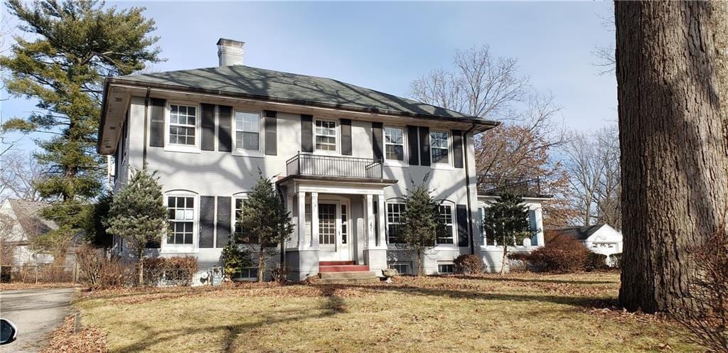 A two-story house is depicted, featuring a symmetrical facade with large windows and a central entrance. Flanking the entrance are manicured shrubs, and a staircase leads to the wide front porch. The surrounding lawn is grassy, with leafless trees casting shadows on the ground.