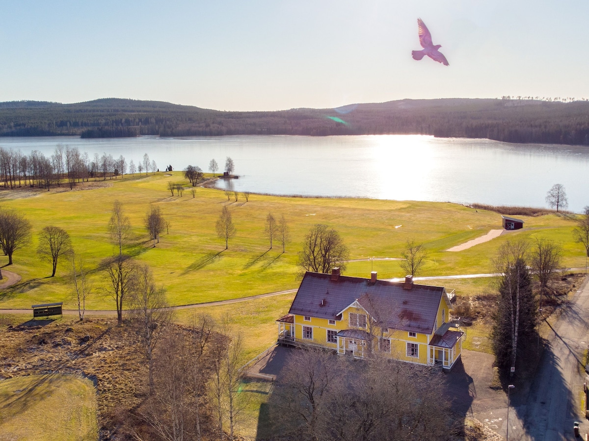 An aerial view captures the spacious yellow building beside a vast green landscape and shimmering Storgösken Lake. The sun reflects on the water's surface, while nearby trees dot the rolling terrain. A golf course can be seen in the foreground, enhancing the natural beauty.