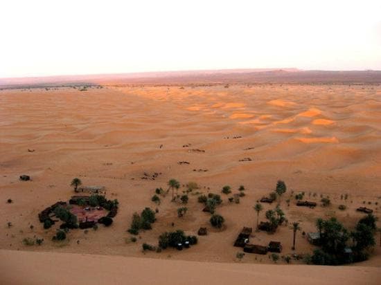 A panoramic view of the vast desert landscape showcases rolling sand dunes in warm golden tones. In the foreground, several structures are nestled among palm trees, indicating a desert retreat. The horizon features a soft gradient of colors as the sun sets, creating a serene atmosphere.