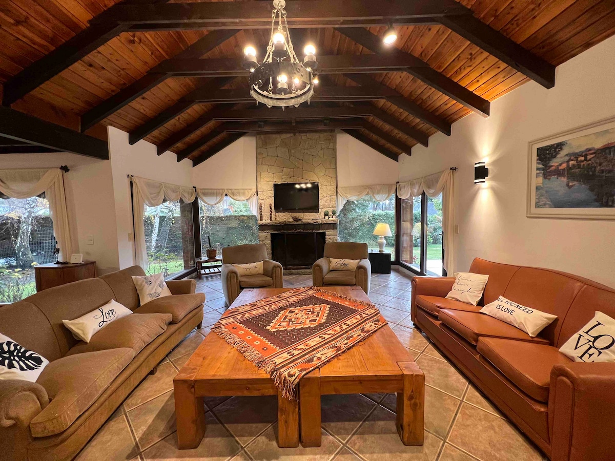 A spacious living area features a vaulted wooden ceiling and natural stone fireplace. Generously sized seating options are arranged around a large wooden coffee table, adorned with a decorative textile. Large windows provide views of the garden, enhancing the room's connection to the outdoors.
