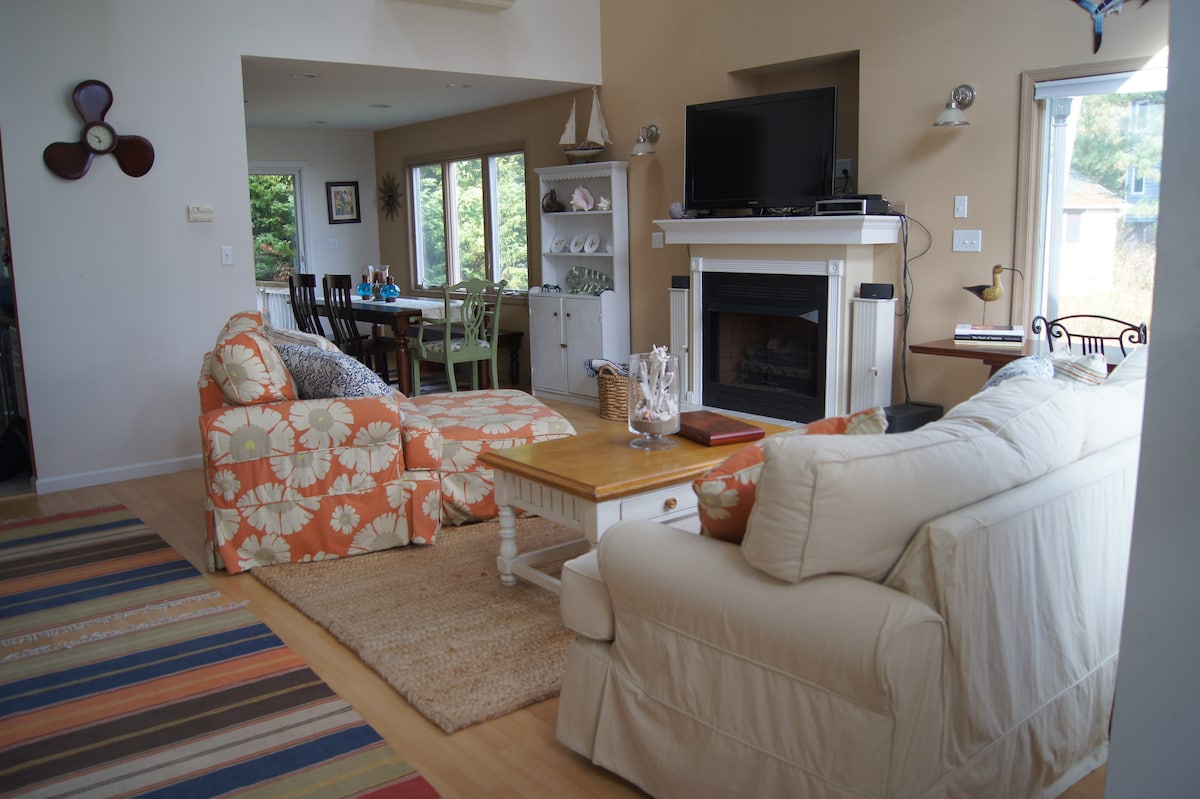 A comfortable living area features a sectional sofa with floral patterns and a cream-colored couch. Natural light fills the space through large windows. A wooden coffee table is centered in front of a fireplace, with a television mounted above. A dining area is visible in the background.
