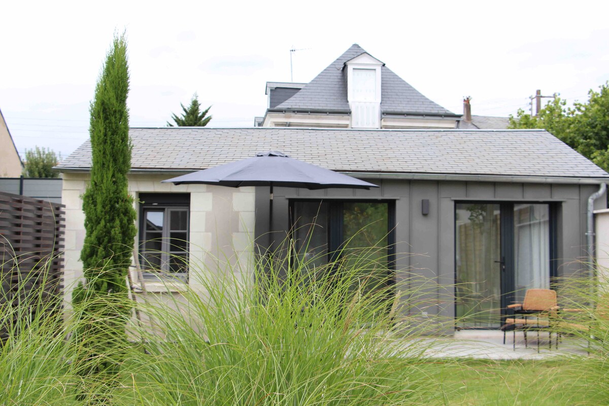 A single-story dwelling is set amidst lush greenery, featuring a gray exterior and large glass doors. A patio area is visible with an umbrella and a chair, providing a shaded seating option. The structure's rooftop is punctuated by a dormer window.