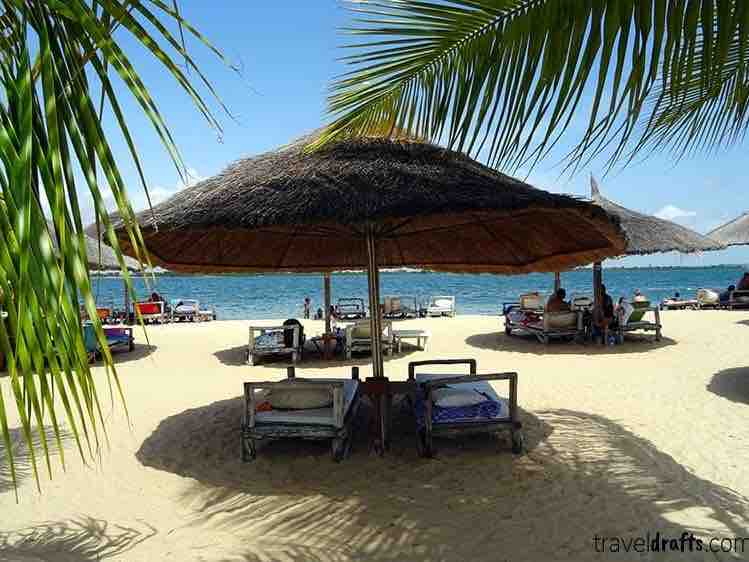 A sandy beach is lined with thatched umbrellas providing shade for sun loungers. Several lounge chairs are arranged around the umbrellas, overlooking the calm sea. Palm fronds are visible, swaying gently in the breeze against a clear blue sky.