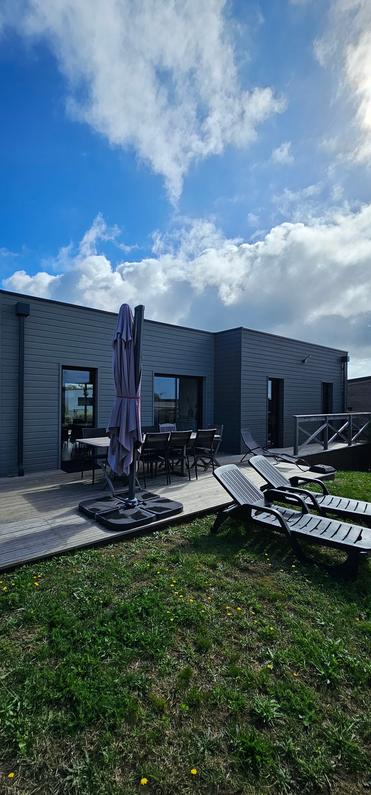 A modern exterior showcases a lovely terrace equipped with lounge chairs and an umbrella. Large windows reflect a serene sky, and a dining set is positioned for outdoor meals. The surrounding grass area features dandelions, contributing to a relaxed outdoor environment.