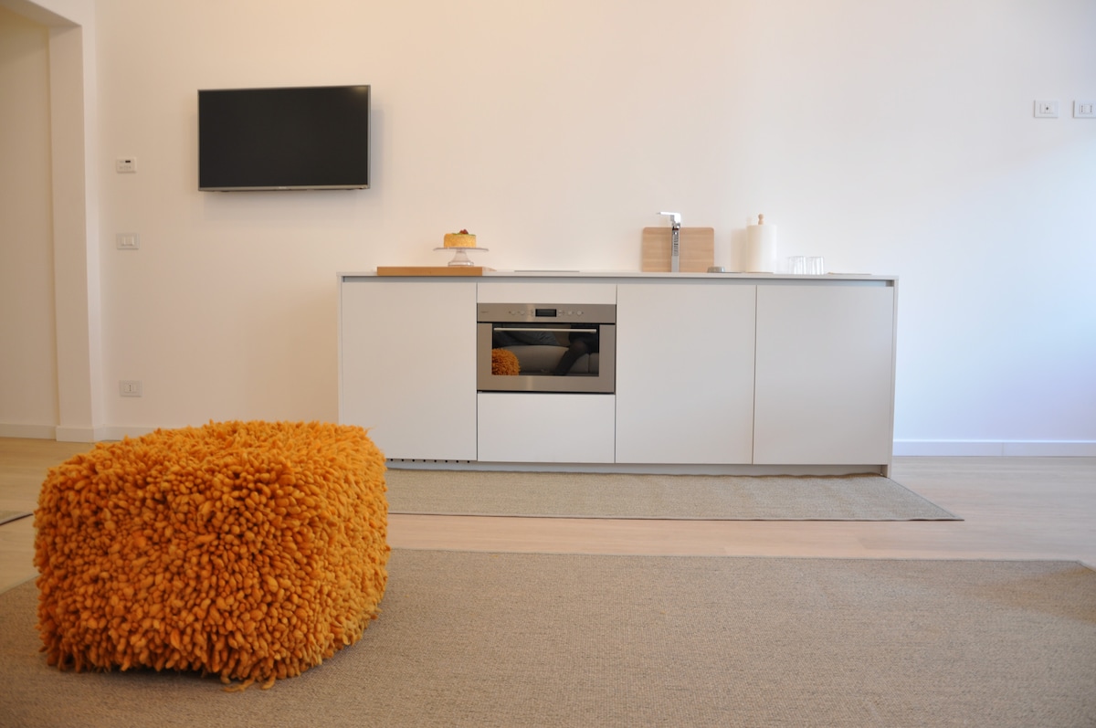 A modern kitchen setup is highlighted with minimalist cabinetry and a built-in oven. A wall-mounted television is visible above the countertop. An orange textured ottoman adds a pop of color to the neutral-toned carpet, creating a cozy seating area.