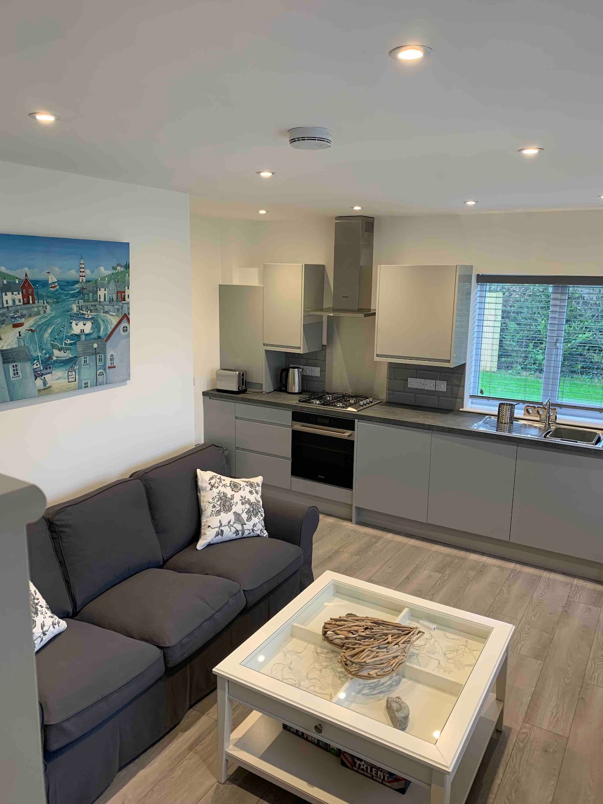 A modern open-plan lounge and kitchen area is depicted, featuring a comfortable gray sofa with decorative cushions. A light-colored coffee table is positioned in front, while a window provides views to the outside. The kitchen includes contemporary appliances and cabinetry.