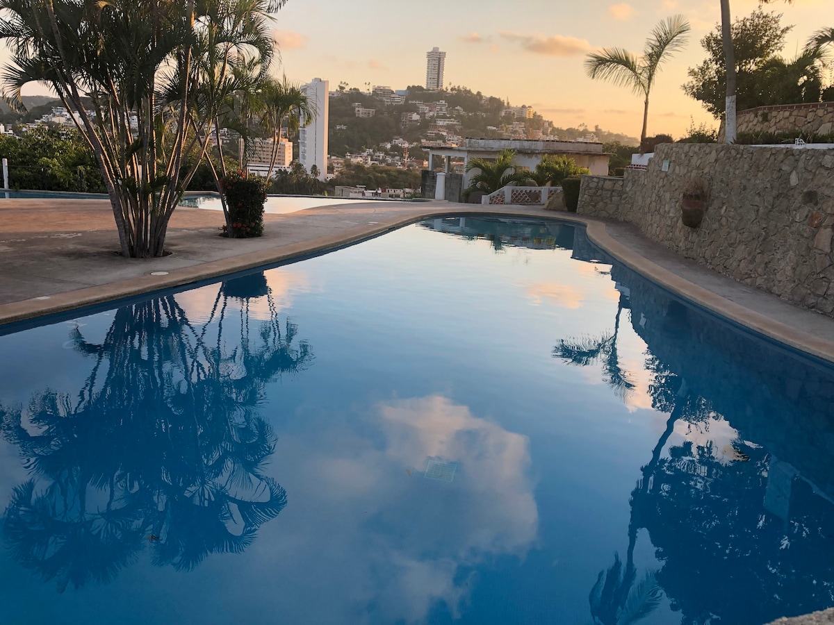 A serene swimming pool reflects the sky and surrounding trees, with an urban backdrop of Acapulco's skyline. The pool area features smooth, paved edges and is surrounded by landscaped plants, providing a tranquil atmosphere for relaxation.