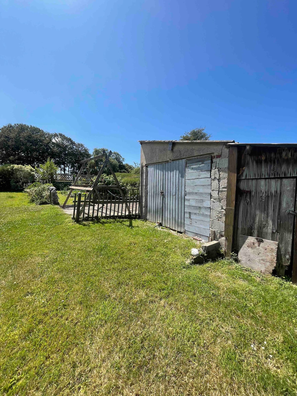 A well-maintained garden area features a grassy lawn under a clear blue sky. A wooden swing set is positioned nearby a rustic shed with a weathered wooden door, set against a backdrop of lush greenery that surrounds the space.