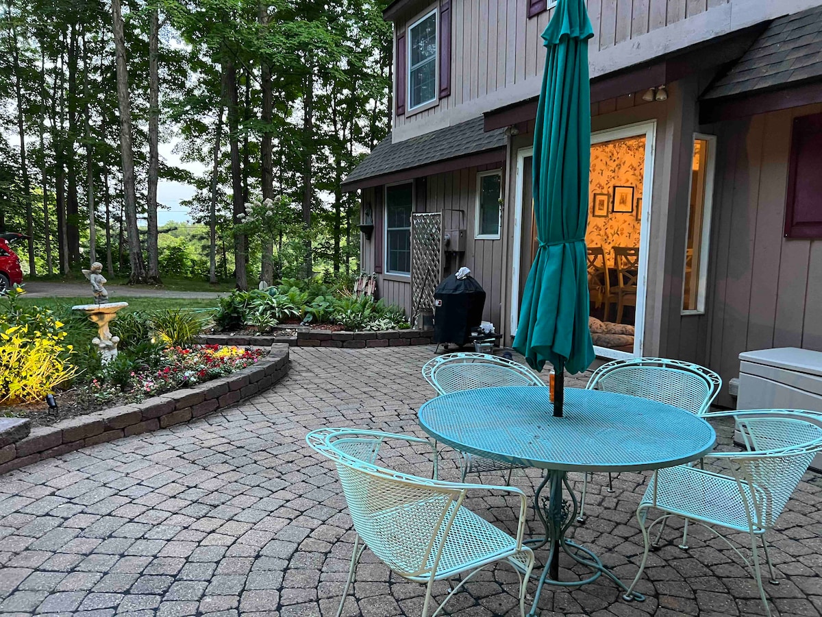 A patio area is seen, featuring a round table surrounded by four light blue chairs. A green umbrella provides shade. The space is bordered by a well-tended garden with colorful flowers and lush greenery. A portion of the house and a scenic view are visible in the background.