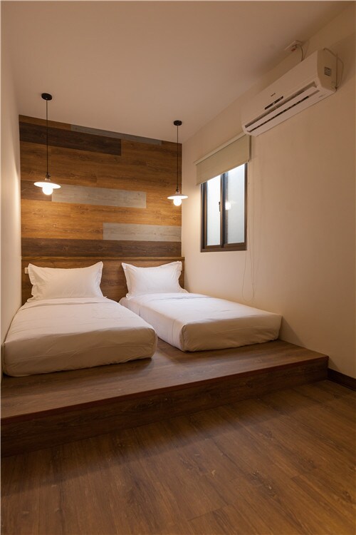 A well-lit sleeping area features two twin beds positioned on a raised wooden platform. Soft white bedding complements the warm wooden wall behind. A small window allows natural light to fill the space, while contemporary light fixtures hang overhead.