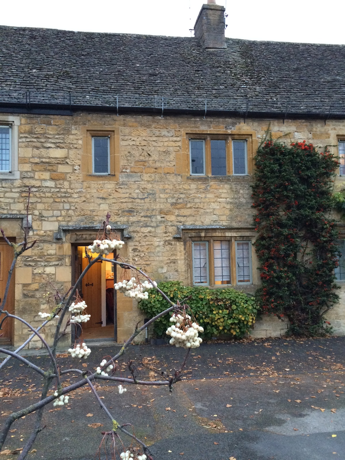 A traditional Cotswold cottage exterior is visible, featuring honey-colored stone walls highlighted by wooden windows and doors. A climbing plant adds a touch of greenery to the entrance. The pathway is lined with fallen leaves, suggesting a tranquil setting at dusk.