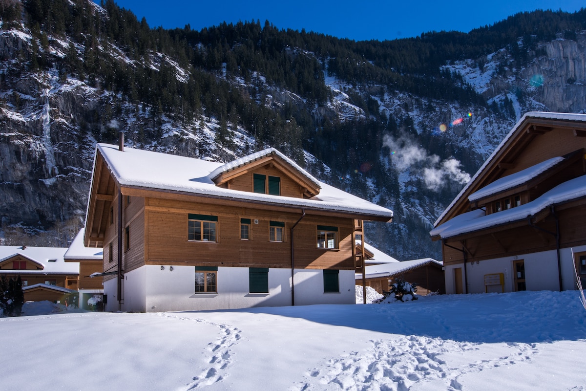 A charming wooden chalet is set against a backdrop of snow-covered mountains. The two-story structure features a combination of wooden and white exterior, with green shutters framing the windows. A blanket of snow covers the ground, creating a serene winter landscape.