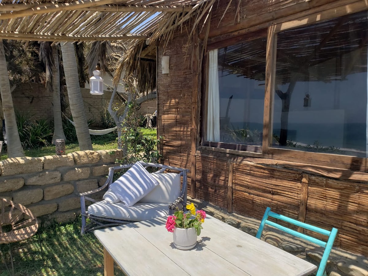 A rustic outdoor seating area is displayed, featuring a wooden table and a comfortable sofa adorned with soft cushions. A potted plant with colorful flowers sits on the table, while palm trees surround the space, creating a serene atmosphere with views of the sea.