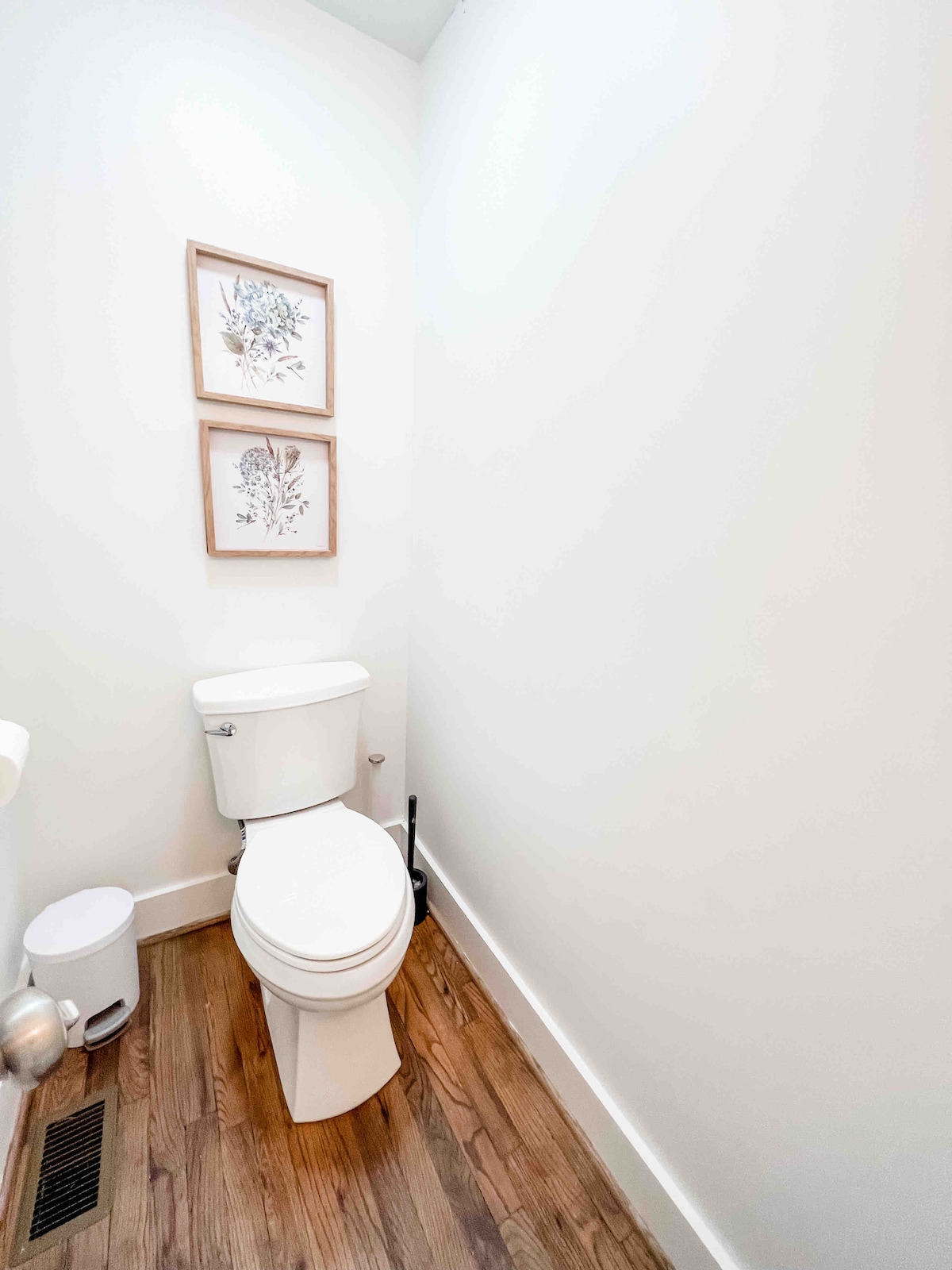 A clean and functional half bathroom is showcased, featuring a white toilet and a small waste bin. The hardwood floor is visible, complementing the minimalistic decor. Two framed botanical prints are hung on a bright wall, adding a touch of interest to the space.