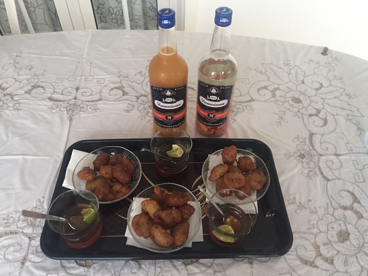 A black tray holds several dishes of accras, a popular local snack, alongside small glasses filled with citrus drinks. Two bottles of beverage are prominently displayed in the background, adding to the presentation. The setting features a light-colored tablecloth with floral patterns.