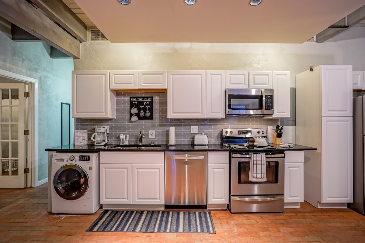 An open kitchen area features white cabinetry and sleek black countertops. Appliances include a microwave, oven, and dishwasher, complemented by a washer/dryer unit. Soft lighting enhances the space, while a striped rug adds an element of warmth to the terracotta flooring.