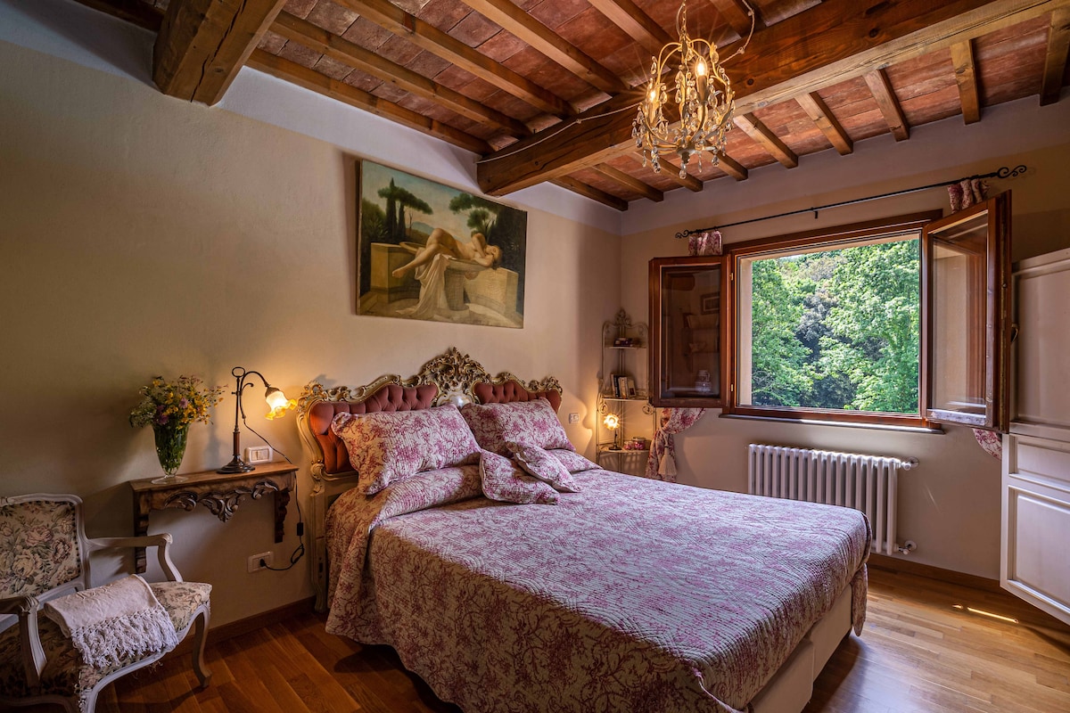 A spacious bedroom features a large floral-patterned bedspread with matching pillows. Natural light enters through a window framed by open wooden shutters, offering a view of the surrounding greenery. A chandelier hangs from the wooden ceiling, and decorative elements include a framed painting and vintage chairs.
