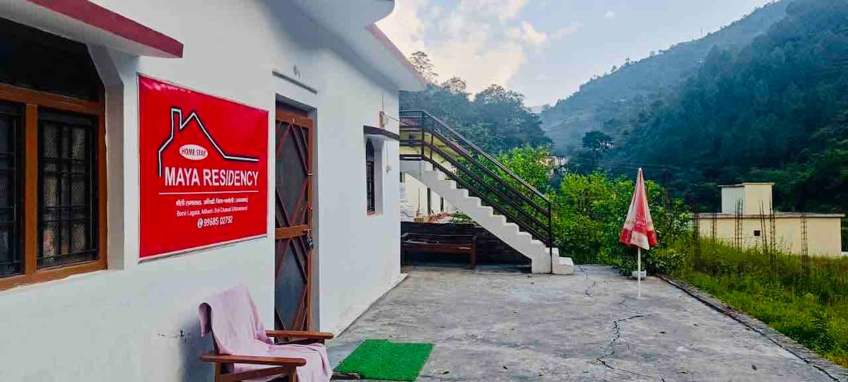A peaceful outdoor space features a concrete terrace with views of lush hills. A wooden sign displays the property name, while a set of stairs leads to the upper level. Nearby, a small area is adorned with seating and greenery.