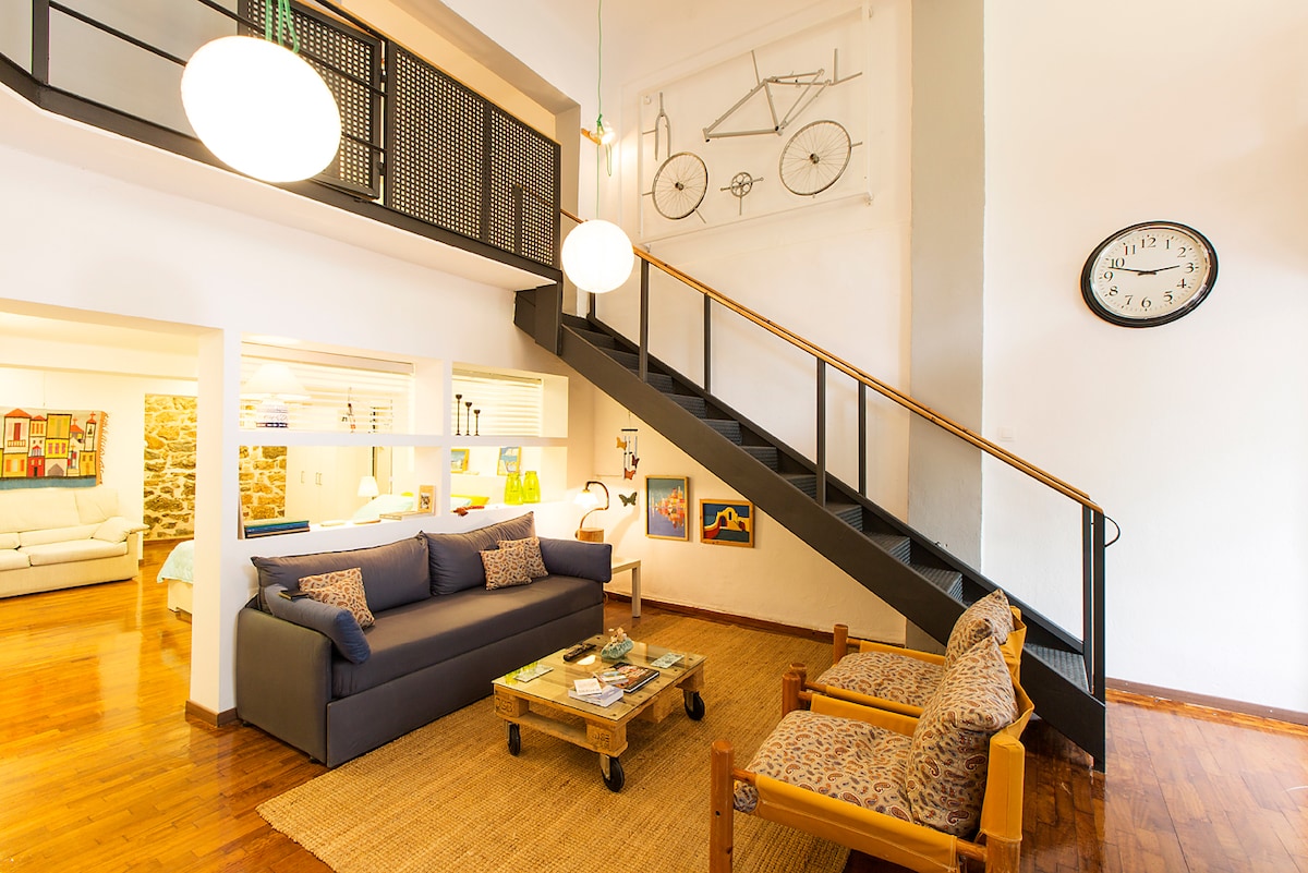 A spacious living area features wood flooring and a high ceiling. A navy sofa is positioned with decorative cushions, complemented by two patterned armchairs and a wooden coffee table. A staircase leads to an upper level. A large wall clock and artwork add visual interest.