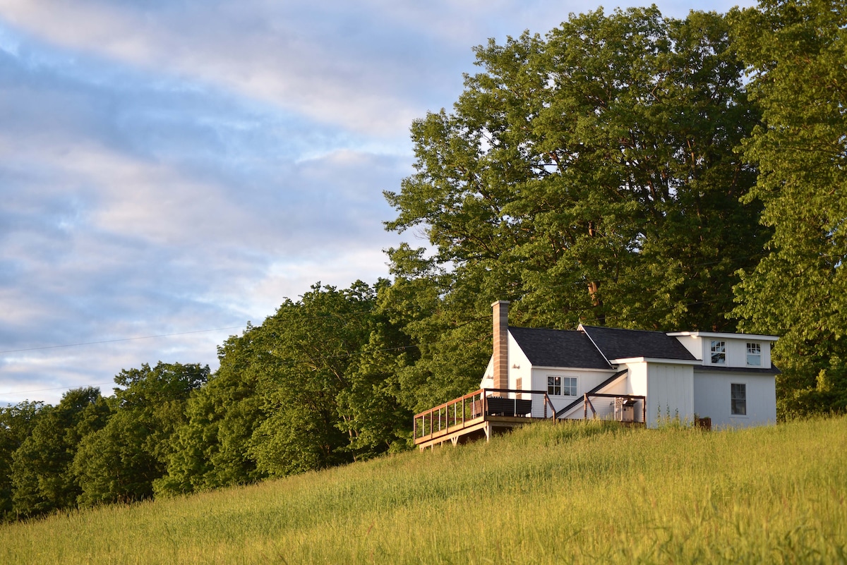 A historic schoolhouse is nestled on a grassy hillside, surrounded by lush greenery. The structure features a prominent deck that extends outward. Soft clouds are visible in the sky, enhancing the serene country setting.