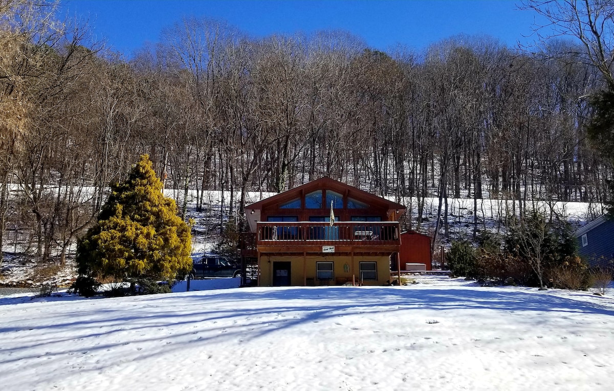 A two-story cottage is framed by a snowy landscape, showcasing a large deck above its entryway. The surrounding area is blanketed in white snow, with leafless trees in the background creating a serene winter scene.