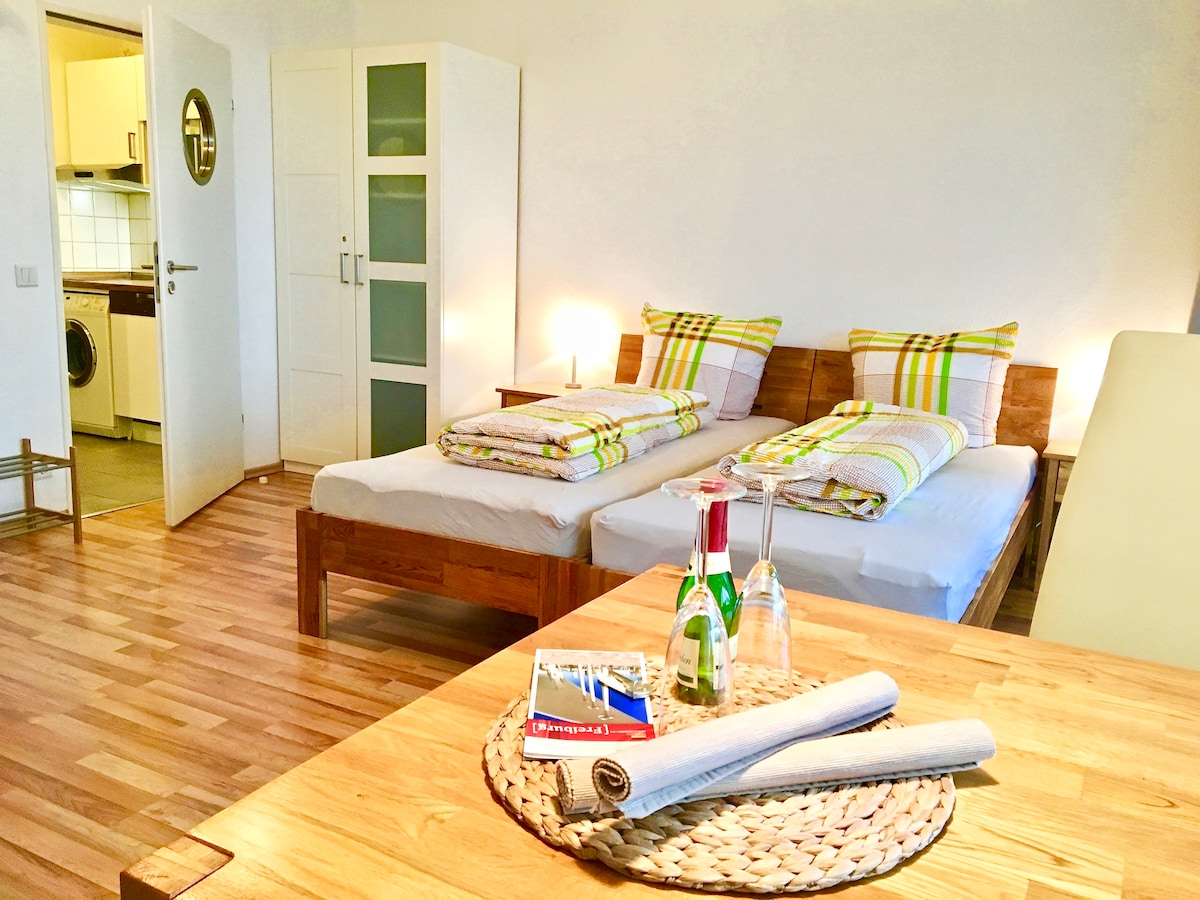 The living area features two spacious beds arranged separately, adorned with patterned bedding. A wooden table with a woven placemat and bottled water is positioned in the foreground. The room is illuminated by soft lighting and includes a glimpse of the kitchen area in the background.