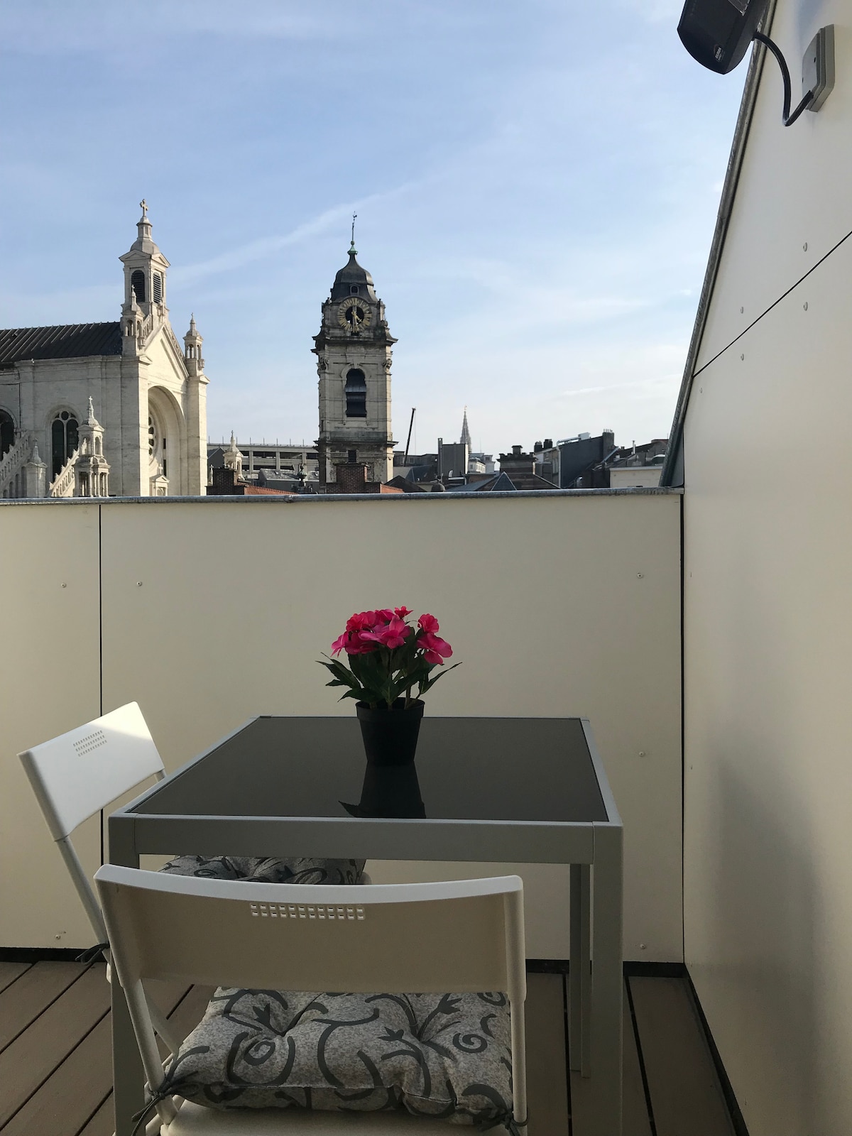 A small balcony features a table and two chairs, set against a backdrop of rooftops and the steeple of the Sainte-Catherine church. A potted flowering plant adorns the table, adding a touch of color to the serene outdoor space.