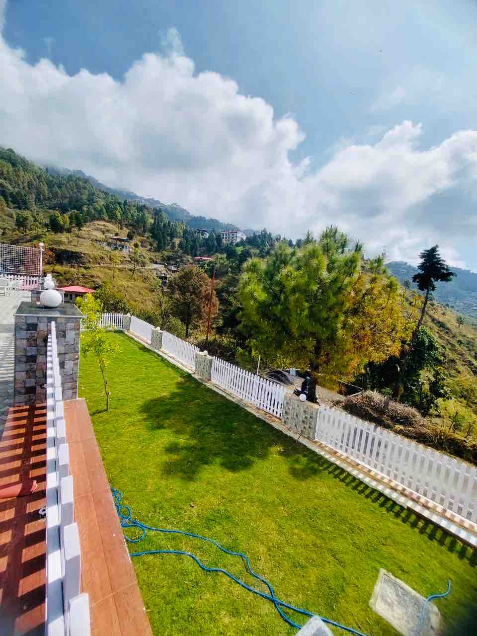 A lush green lawn is bordered by a white picket fence, offering expansive views of the surrounding hills and trees. The sky is bright with fluffy clouds, and sunlight highlights the natural landscape, creating an inviting outdoor space for relaxation.