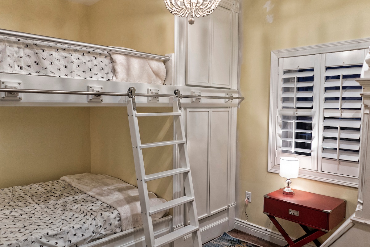 A private suite features custom queen bunk beds built into white cabinetry, with a spacious lower bed and a ladder leading to the upper bunk. The room’s yellow walls complement the wooden accents. A small nightstand with a lamp and a window with plantation shutters are visible.