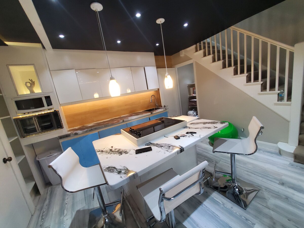 A modern kitchen features a stylish marble-top island with seating, illuminated by pendant lights. Sleek cabinetry, equipped with appliances, lines the wall, while a staircase leads to an upper level. The space is designed with a blend of light and dark tones.