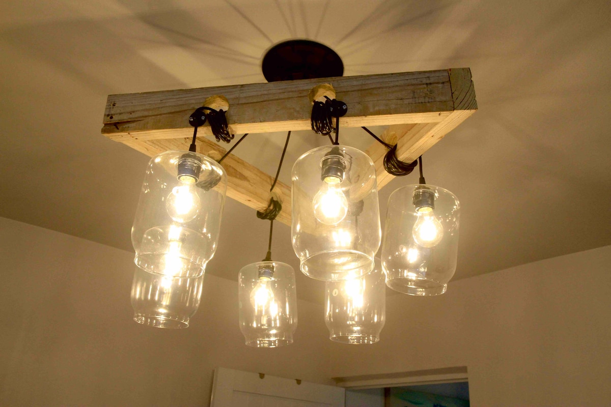 A unique light fixture features a wooden frame holding five glass jar bulbs, providing ample illumination. The arrangement creates a warm glow, with each bulb suspended by black cords that enhance the industrial aesthetic of the space.