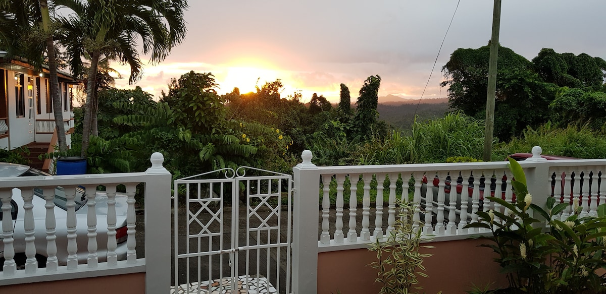 A view of the sunset is framed by a decorative white gate, with lush greenery surrounding the property. The horizon showcases vibrant colors as sunlight filters through the trees, creating a serene backdrop. The entrance features a tastefully designed fence with decorative posts.