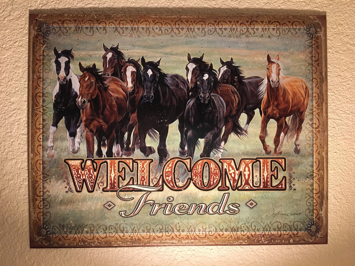 A rustic welcome sign displays the phrase 'WELCOME Friends' in bold letters. The background features a group of horses, varying in colors from dark brown to chestnut, standing together in a grassy field. The artistic design captures the essence of the ranch setting.
