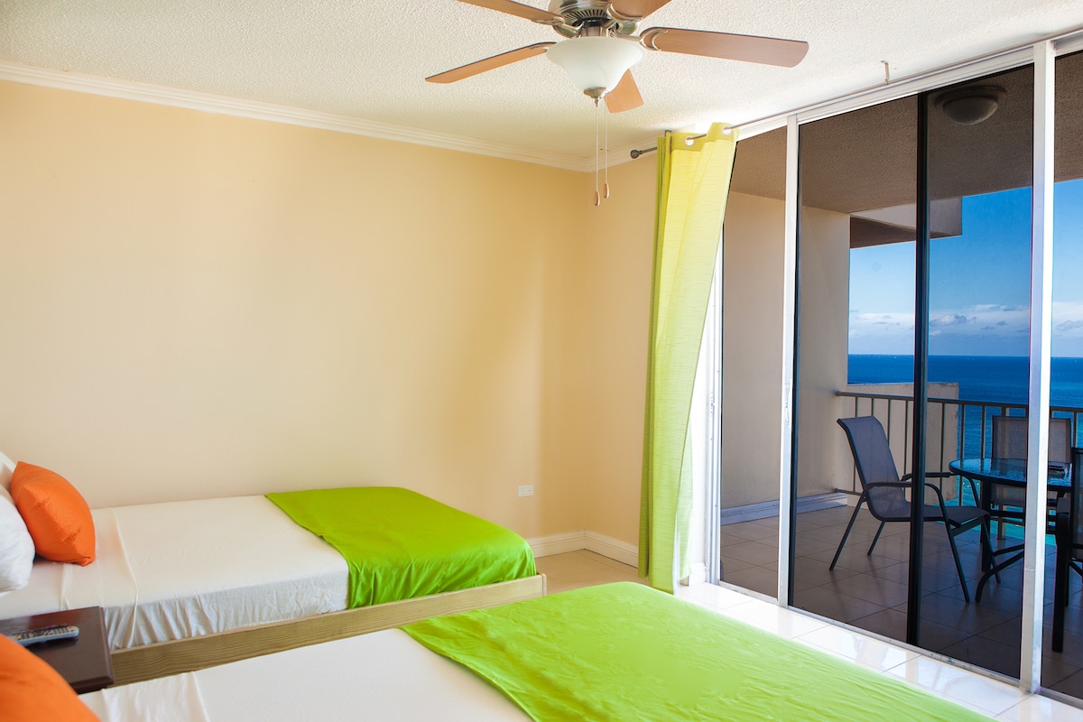 A light-filled second bedroom features two full-size beds adorned with vibrant green linens. A ceiling fan creates airflow in the space. Large glass doors open to an ocean view balcony, allowing natural light to illuminate the room.