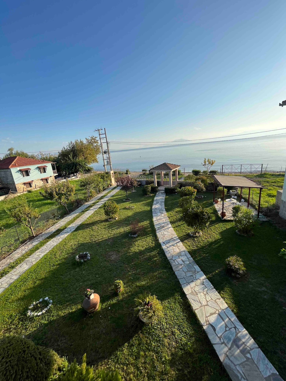 A well-maintained outdoor space is visible, featuring neatly arranged stone pathways that wind through lush greenery. Several shrubs and small trees are present, along with a gazebo and shaded area. The sea can be seen in the background, enhancing the serene atmosphere of the garden.