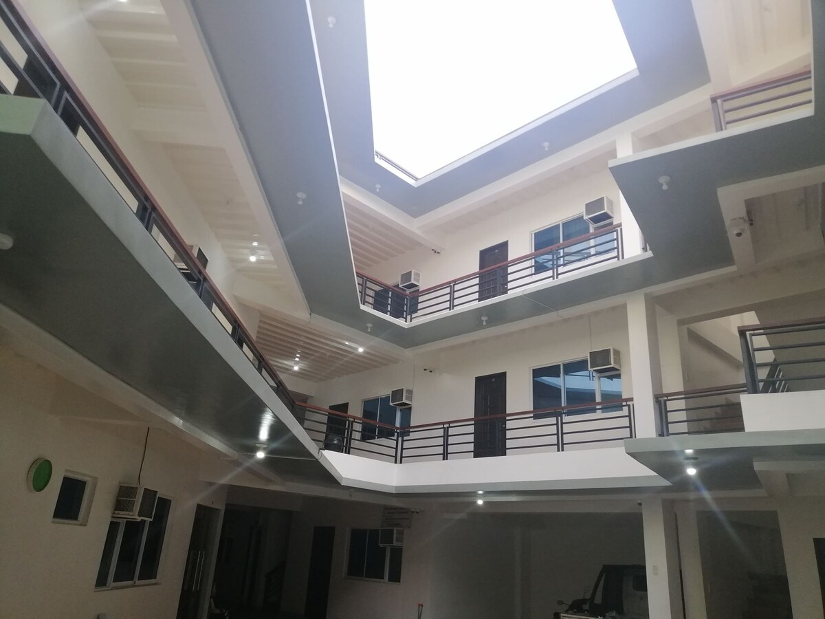 An open atrium is visible, featuring multiple levels with air conditioning units mounted on the walls. Skylights provide natural light, illuminating the space. Surrounding balconies can be seen, with doors leading into the rooms, creating a sense of openness and accessibility.