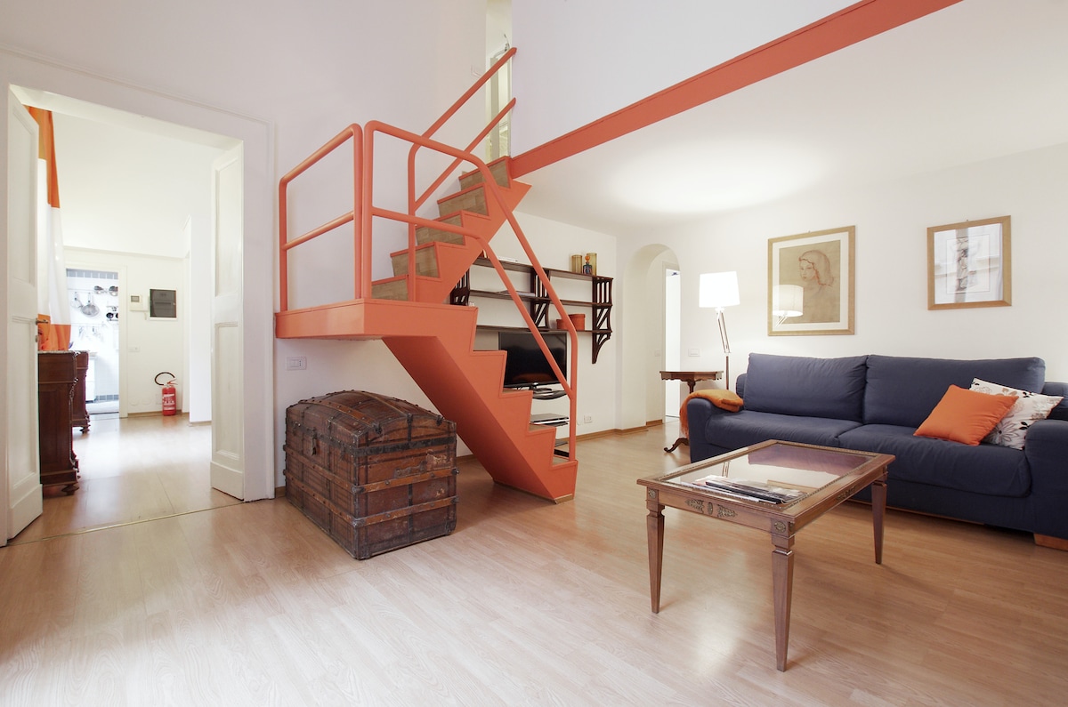 A spacious living area features a comfortable blue sofa with orange accents. A wooden coffee table sits in front, and a vintage trunk is positioned beside the stairs. The staircase, with an orange railing, leads to a second level, while artwork is displayed on the walls.