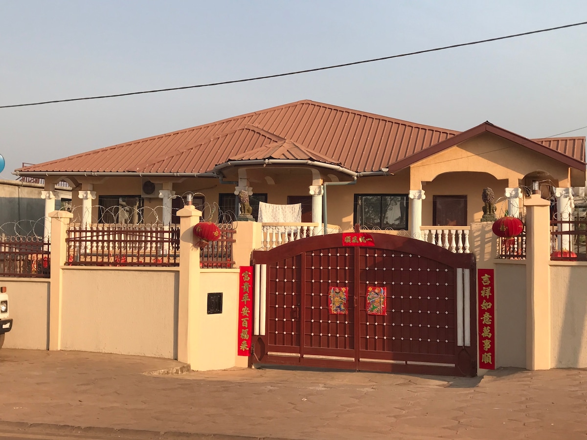 The exterior of the house features a welcoming gated entrance, adorned with traditional lanterns. The structure showcases a spacious porch area with seating, complemented by a distinctive roof design. A well-maintained garden area is visible, enhancing the overall inviting appearance.