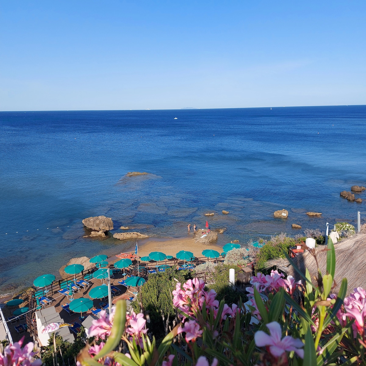A view of clear blue waters extends towards the horizon, with distant boats visible. Below, a rocky shoreline meets a sandy beach, where individuals can be seen enjoying the surroundings. Lush greenery and blooming flowers frame the scene, adding natural beauty.