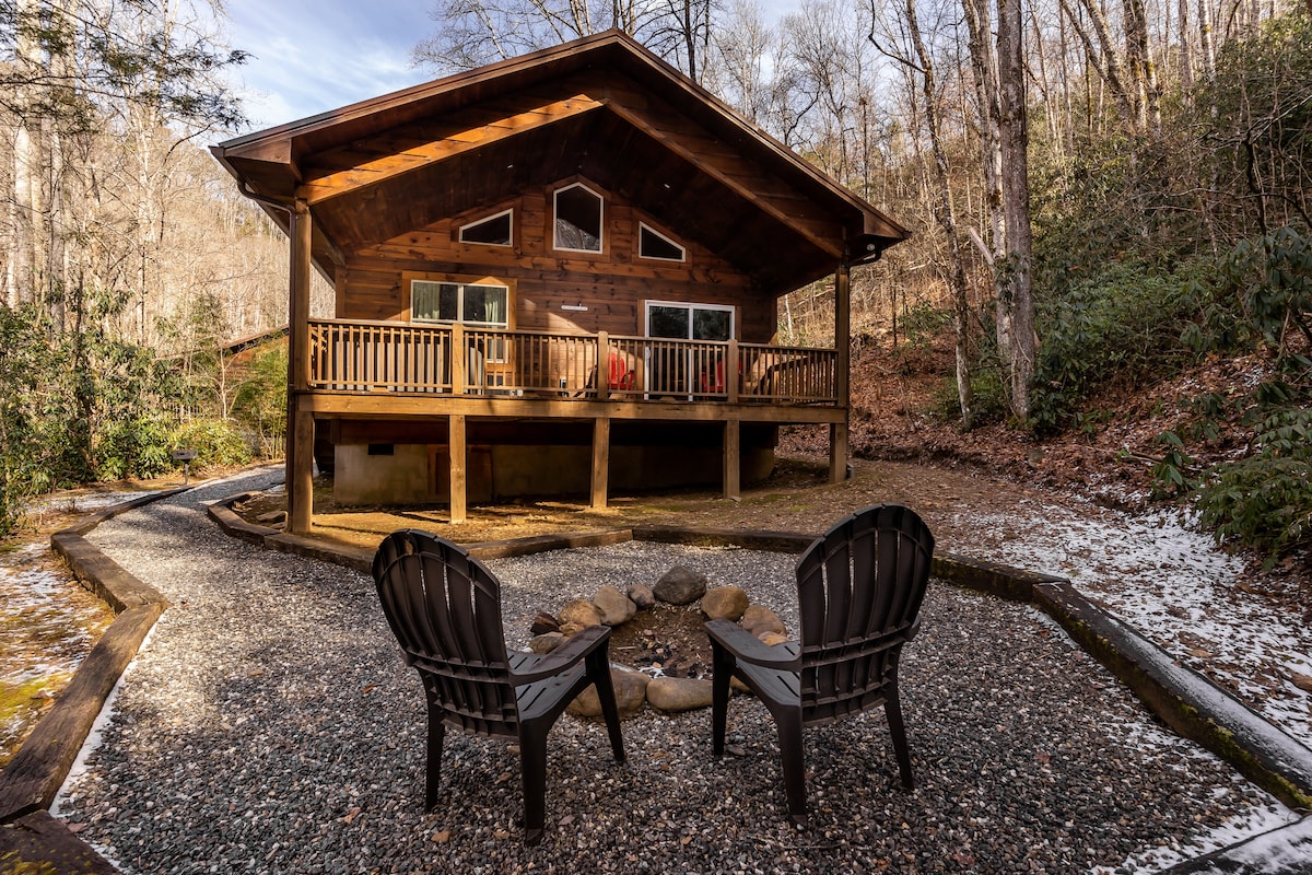 A beautiful log cabin is nestled among trees, featuring a spacious covered deck with railings, perfect for enjoying nature. A fire pit area with two black chairs is set in the foreground, surrounded by gravel, offering a cozy spot for gatherings.