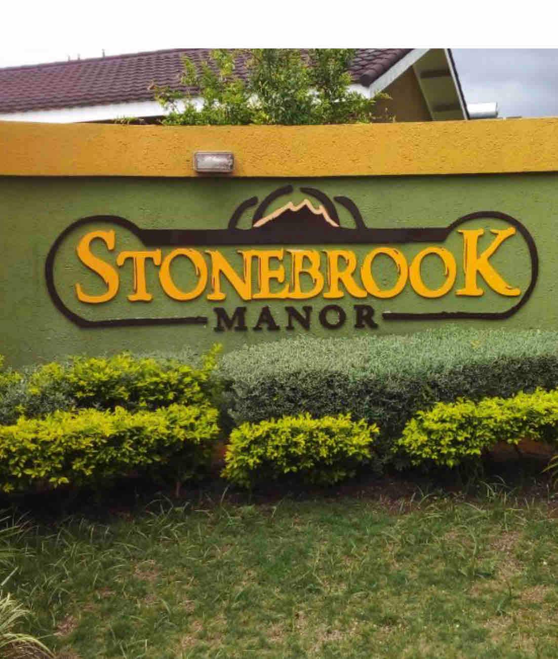The entrance sign of Stonebrook Manor is prominently displayed, featuring bold yellow lettering against a green background. Lush, manicured shrubs and plants frame the sign, enhancing the welcoming feel of the property.