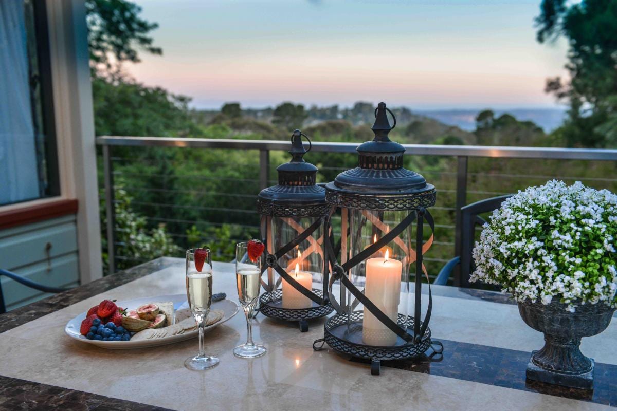 Two elegant lanterns are positioned beside glasses of champagne on a marble table, accompanied by a plate of assorted fruits and pastries. A lush green landscape is visible in the background, with the soft hues of twilight creating a serene atmosphere.