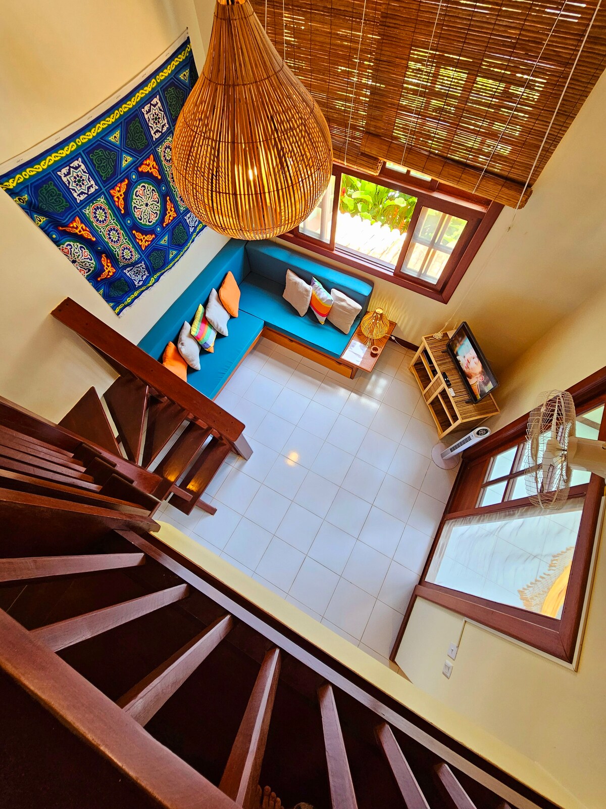 A view from above captures the living area with a bright blue sofa adorned with colorful cushions. Natural light flows through large windows, highlighting a television unit and a fan. The staircase, featuring wooden railings, leads to the upper level.